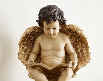 May include: A golden cherub statue with dark hair and feathered wings, reading a book while seated on a golden sphere. A book with the text "Lest We Forget" and a candle are nearby. Decorative home decor.