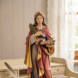 May include: A detailed statue of a woman in a flowing gown, holding a book and a palm frond. The statue has a red and gold cloak and a crown. The statue is on a wooden base, and is approximately 12 inches tall.