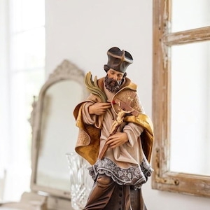 May include: A hand-painted wooden statue of a bearded figure wearing a dark cap, a tan robe with gold trim, and a white lace-trimmed garment. The figure holds a wooden crucifix and a palm frond while standing on an open book atop a dark base.