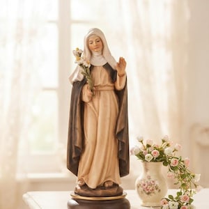 May include: A hand-painted wooden statue of a nun wearing a white head covering and a long, flowing beige habit with a dark brown cloak. The figure holds a cluster of white lilies in one hand and has the other hand raised in a gesture of blessing.