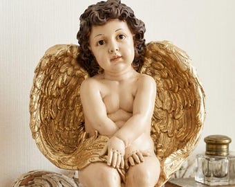 May include: A detailed statue of a cherub with golden wings, seated and looking forward. The cherub has brown curly hair and is nude. The wings are detailed and ornate. The statue is on a distressed white table.