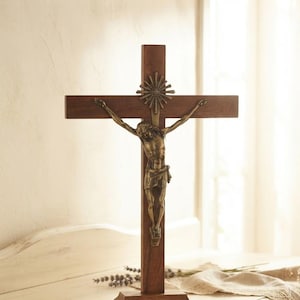 Può includere: Un crocifisso in legno da appoggio con una croce marrone scuro su base rettangolare e una figura metallica color bronzo di Gesù fissata al centro. Sopra la figura, un piccolo emblema a raggiera è fissato sulla parte superiore della croce.