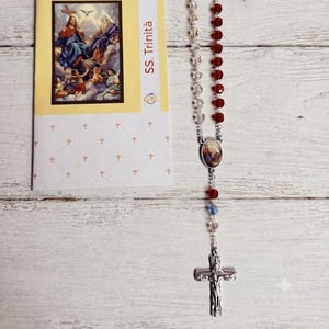 May include: A religious rosary featuring blue, red, and clear beads with a silver-toned crucifix and a central medal, accompanied by a rectangular prayer card with the text SS. Trinità and an illustration of the Holy Trinity.