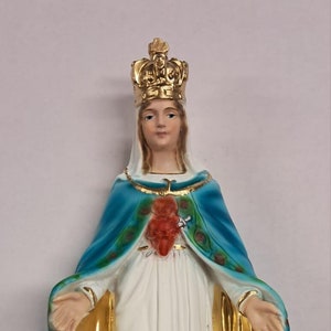 May include: A religious figurine of a woman with a gold crown, wearing a white dress and a blue cloak with gold trim. A red heart is visible on the chest. The base is light blue with the text "N.D. DU. CAP."