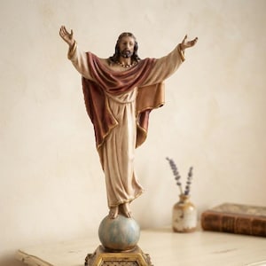 Può includere: Una statuetta raffigurante una figura barbuta con le braccia tese, che indossa una veste color crema con un drappeggio bordeaux sulle spalle. La figura poggia su una sfera dai toni blu, montata su una base quadrata dorata e decorata.