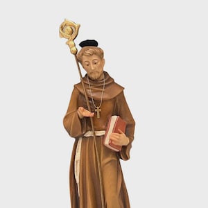 Pode incluir: Estátua castanha de uma figura religiosa segurando um cajado com um topo dourado, um livro vermelho e um colar com uma cruz. A figura está vestida com uma túnica castanha e um chapéu preto. A estátua está sobre uma base retangular.