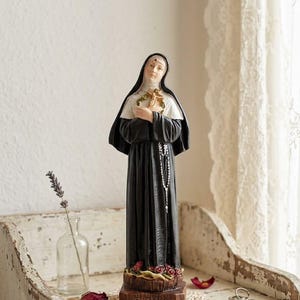 May include: A standing figurine of Saint Rita wearing a black and white habit, holding a small wooden crucifix and a crown of thorns. The base is brown with the text S. RITA engraved on the front, decorated with small red roses at the feet.