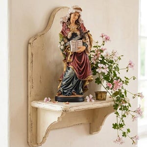 Może przedstawiać: Religijna statua kobiety w długiej sukni, trzymającej książkę i miecz, stoi na postarzałej białej półce. Statua jest ozdobiona złotymi akcentami i otoczona różowymi kwiatami. Półka jest zamontowana na ścianie.