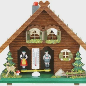 May include: A wooden chalet-style weather station with a pitched roof, featuring miniature figures in traditional attire. The chalet has a thermometer, decorative trees, and a deer figurine. The color palette includes shades of brown, green, red, and white.
