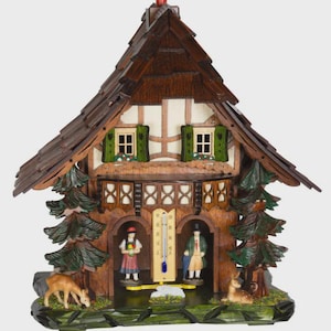 May include: A wooden cuckoo clock in the shape of a chalet. The clock features a dark brown roof, white walls, and green shutters. Inside are miniature figures and a thermometer. Two deer and two pine trees are on the base.