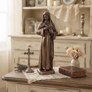May include: A bronze-toned statue of a cloaked figure holding a small crucifix, standing on a rectangular base. The figure wears a long, flowing robe with a hood and holds a rosary in one hand, capturing a serene and solemn expression.