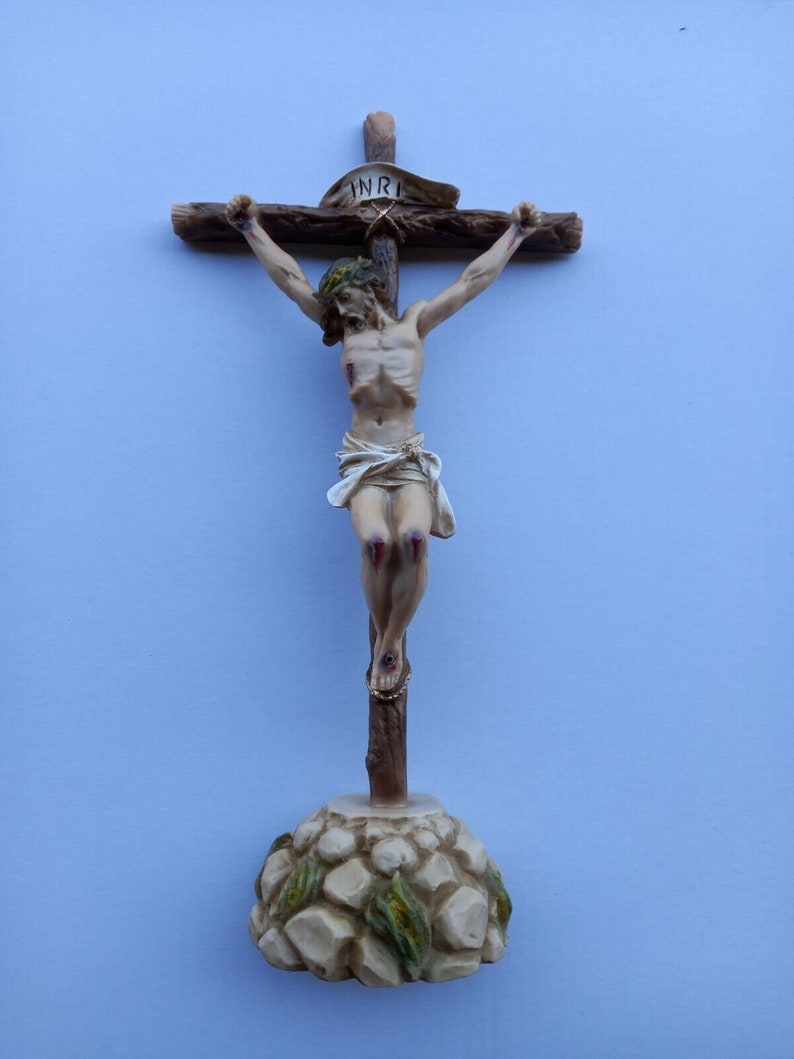 May include: A wooden crucifix with a sculpted figure of Jesus Christ on the cross. The figure is painted in a realistic style with details of wounds and blood. The cross is mounted on a base that resembles a rocky hill.