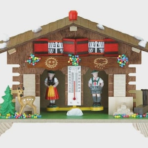 May include: A wooden chalet-style thermometer with a brown roof, featuring miniature figures in traditional attire. The chalet has a thermometer with Fahrenheit and Celsius scales, and decorative elements like a deer and a small tree.