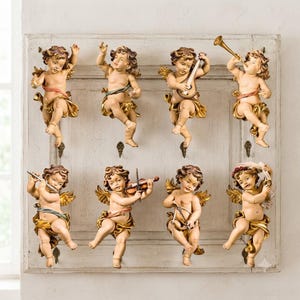 Pode incluir: Um painel de parede retangular de madeira branca desgastada com oito figuras de querubins esculpidas em duas filas. Cada querubim tem asas douradas e drapeados coloridos, sendo que alguns seguram instrumentos musicais como trompete ou violino.