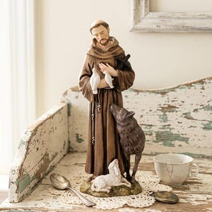 May include: A standing figurine of a monk wearing a brown hooded robe, holding two white doves and a black cross. A dark brown wolf stands beside the monk, while a small white lamb rests at the base on a textured, earth-toned pedestal.