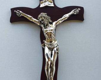 May include: A silver-colored Jesus figure on a dark brown wooden cross. The figure is depicted with outstretched arms and a crown of thorns. The cross has a small plaque with the inscription "INRI" at the top.