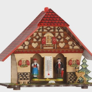 May include: A wooden weather house with a red roof and decorative cutouts. The house features miniature figures of a man and woman, a thermometer, a deer, and a small fir tree. The house is designed with a traditional aesthetic.