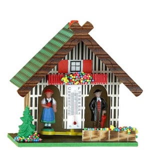 May include: A decorative wooden chalet-style thermometer with a green roof and a small window. It features miniature figurines of a man and woman in traditional attire, and a temperature gauge in Fahrenheit and Celsius.