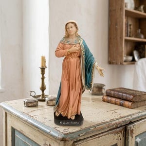 May include: A statue of a woman in a long peach-colored dress and a blue shawl. The statue has a gold-colored belt and trim. The base of the statue is black with the text "N. S. DO PARTO".