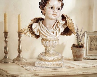 May include: A decorative bust of a cherubic figure with dark brown curly hair, a pale complexion, and gold-toned wings. The figure is draped in a white cloth and rests upon a textured, pedestal-style base with a weathered, off-white and gold finish.