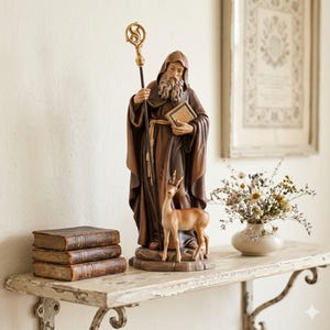 May include: A detailed statue of a robed figure holding a staff and book, with a small deer at its feet. The statue is brown and gold, set on a distressed white shelf with old books and a vase of flowers. The background is a neutral wall.