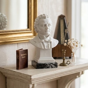 May include: A white marble bust of a man on a black and white base, with the name "SCHUBERT" inscribed. A small brown book with gold lettering, a metronome, and a brass candlestick are also on the shelf.