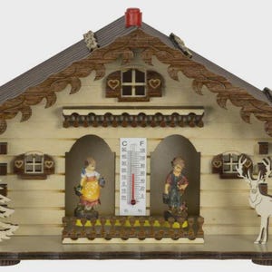 May include: Wooden chalet-style thermometer with a brown roof and decorative trim. The chalet features two figurines, a deer, and a thermometer displaying both Celsius and Fahrenheit. The chalet is painted in light beige and brown.