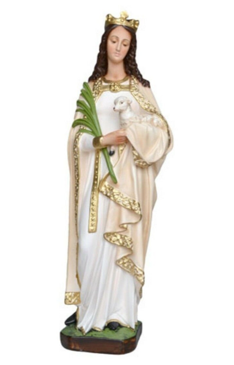 May include: A white statue of a woman wearing a crown and a long white robe with gold trim. She is holding a lamb in her left arm and a branch in her right hand.