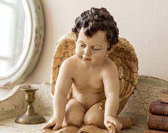 May include: A classical angel figurine with golden wings and a book. The angel is seated, reading a large, open book. The statue has a cream-coloured body and dark brown hair. The wings and book are gold, creating a vintage aesthetic.