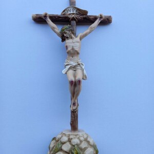 May include: A wooden crucifix with a white and brown Jesus figure. The figure has a crown of thorns and blood stains on its hands and feet. The crucifix has a white banner with the word "INRI" on it. The base of the crucifix is made of stone with green leaves.