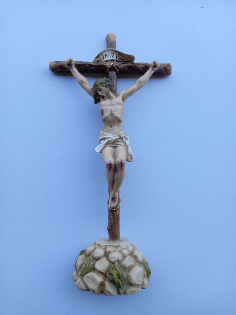 May include: A wooden crucifix with a white and brown Jesus figure. The figure has a crown of thorns and blood stains on its hands and feet. The crucifix has a white banner with the word "INRI" on it. The base of the crucifix is made of stone with green leaves.