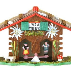 May include: A wooden chalet-style decorative piece with a red roof, brown walls, and green accents. The chalet features two figures in traditional clothing, edelweiss flowers, and a small mushroom. The roof has snow-like details.