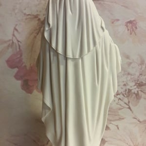 Puede incluir: Figura blanca de la Virgen Mar&iacute;a, de pie con las manos juntas en oraci&oacute;n. La figura lleva una larga t&uacute;nica blanca y un velo blanco. La base de la figura es blanca y tiene las palabras "MADE IN ITALY" inscritas en ella.