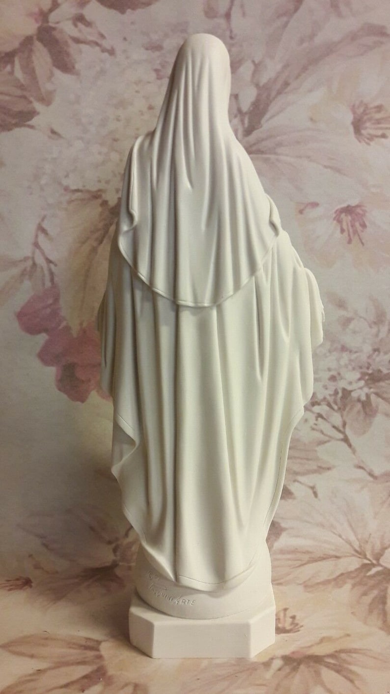 Puede incluir: Figura blanca de la Virgen Mar&iacute;a, de pie con las manos juntas en oraci&oacute;n. La figura lleva una larga t&uacute;nica blanca y un velo blanco. La base de la figura es blanca y tiene las palabras "MADE IN ITALY" inscritas en ella.