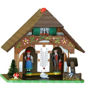 May include: A wooden chalet-style weather station with a thermometer, featuring figurines of a man and woman in traditional attire. The chalet has a brown roof, green shutters, and floral accents. A small tree and mushroom are on the green base.