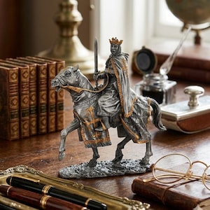 May include: A detailed figurine of a knight on horseback, holding a sword aloft. The knight wears a crown and cape, with gold accents on the armor and horse's tack. The statue is silver and gray, and sits on a wooden surface.