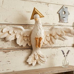 Könnte beinhalten: Eine Wandskulptur einer weißen Taube mit ausgebreiteten Flügeln, die einen goldenen dreieckigen Heiligenschein über dem Kopf und orangefarbene Füße aufweist. Der Vogel ist in Flugposition dargestellt und zeigt detaillierte Federstrukturen.