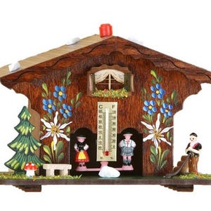 May include: A wooden chalet-style thermometer with painted floral and alpine details. The chalet features a thermometer, miniature figures, and a small fir tree. The roof is light brown, and the walls are dark brown.