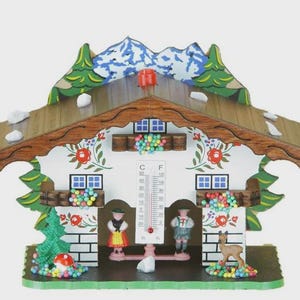 May include: A decorative wooden chalet-style thermometer with a brown roof, white walls, and colorful floral accents. The chalet features miniature figures, a deer, and a thermometer scale in Fahrenheit and Celsius.