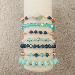 May include: A stack of eight bracelets with a mix of turquoise and dark blue beads, some with rose gold accents. The bracelets are stacked on a white display stand.