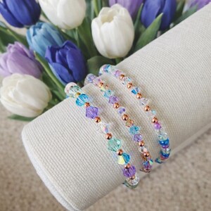 May include: Three beaded bracelets with iridescent, purple, blue, and clear crystals and copper-colored beads are displayed on a white fabric roll. A bouquet of blue, purple, and white tulips is in the background.