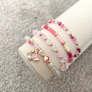 May include: A set of six pink and white beaded bracelets with a gold butterfly charm. The bracelets are stacked on a white surface.