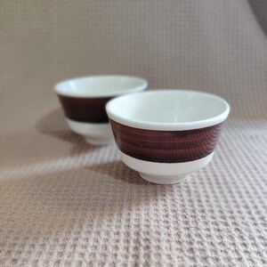 May include: Two small, white ceramic bowls with a dark brown, wood-grain pattern around the top and a white base. The bowls are set against a neutral background, showcasing their simple, yet elegant design.