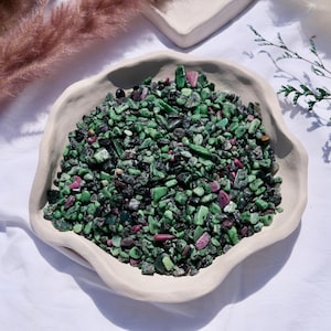 May include: A white ceramic bowl filled with green, black, and pink tumbled stones. The stones are of various sizes and shapes.