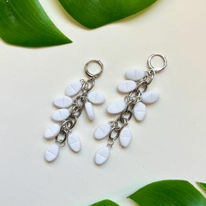 May include: A pair of dangle earrings featuring silver-toned metal chains adorned with multiple small, white, oval-shaped pill charms. Each earring is attached to a circular silver-toned hoop closure, creating a cascading, layered design of white tablets.