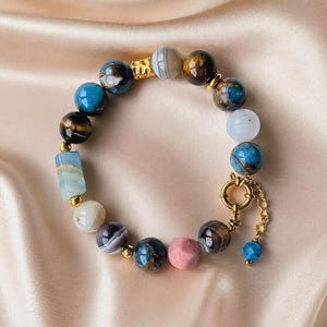 Op de afbeelding: Een kralenarmband met diverse ronde en cilindervormige stenen in blauwe, bruine, witte, grijze en roze tinten. De kralen zijn voorzien van kleine goudkleurige metalen tussenstukjes en een ronde gouden sluiting met een bijbehorend kettinkje.