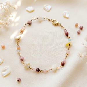 Könnte beinhalten: Ein filigranes, goldfarbenes Armband mit einer Vielzahl von Perlen in Rosa-, Weiß- und Goldtönen. Das Armband umfasst runde, ovale und unregelmäßig geformte Perlen mit einem goldenen Verschluss. Zusätzliche Perlen und Muschelsplitter sind um das Armband verstreut.