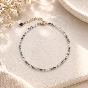 Puede incluir: Una pulsera delicada con pequeñas cuentas facetadas transparentes y gris oscuro dispuestas en un patrón alterno. La pieza incluye una cadena extensora de metal dorado con un cierre de mosquetón y una pequeña cuenta redonda al final.