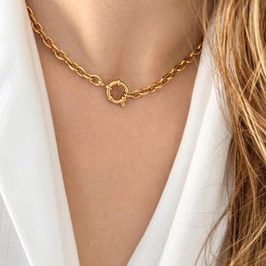 Peut inclure: Un collier chaîne doré avec un grand fermoir. Le collier est composé de maillons épais de forme ovale. Le fermoir est de forme circulaire avec un petit levier. Le collier est porté sur une chemise blanche. Un bijou élégant.