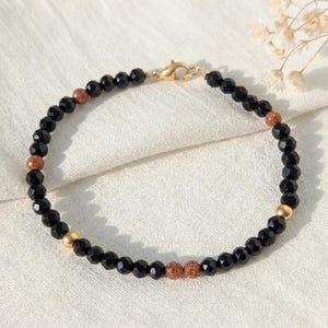 Puede incluir: Una pulsera de cuentas negras facetadas, intercaladas con cuentas doradas y color bronce. La pulsera se asegura con un cierre dorado. La joya se exhibe sobre una superficie blanca texturizada.